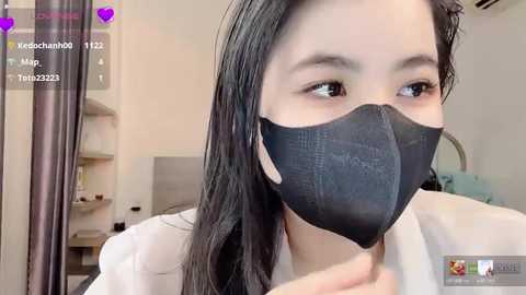 Video of an East Asian woman with long black hair, wearing a black face mask, white top, and glasses, in a sterile medical setting.