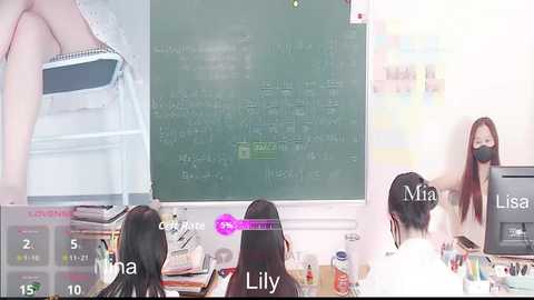 Video of three Asian women in a classroom. One sits at a desk, two stand by a chalkboard, wearing face masks. Background shows classroom clutter.