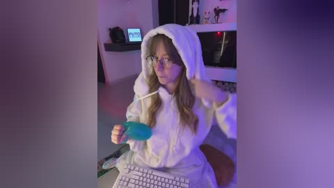 Video of a young woman in a white, fuzzy, hooded robe, sitting at a desk with a computer, holding a blue marker. Background features a monitor displaying a video game.