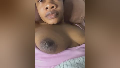 Video of a Black woman lying on her back, partially covered by a pink sheet, showing her bare breast with a prominent, dark areola and nipple.