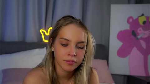 A video of a young blonde woman with light skin and a small nose, wearing minimal makeup, sitting in a bedroom with pink and white decor, and a neon \"V\" sign.