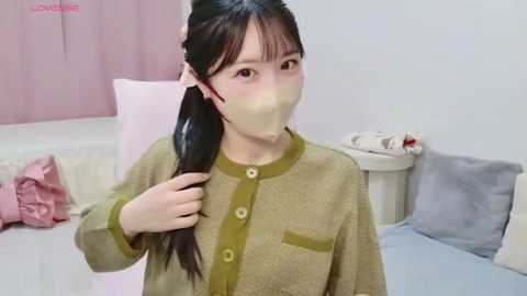 Video of an East Asian woman with long black hair, wearing a green, textured cardigan, and a beige face mask, adjusting her ponytail in a pastel-toned bedroom.