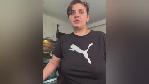 Video of a woman with short brown hair, wearing a black Puma t-shirt, in a modern kitchen with white cabinets and a tiled backsplash.