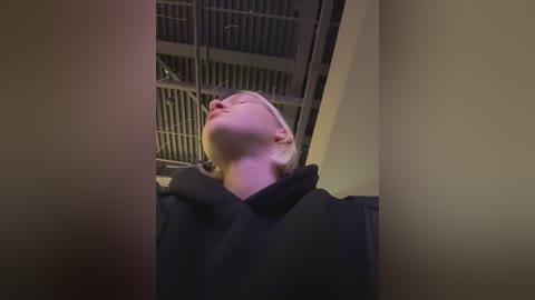 Video of a blonde woman in a black coat, tilted back in an elevator, eyes closed, looking up. Background shows metallic elevator door and dim lighting.