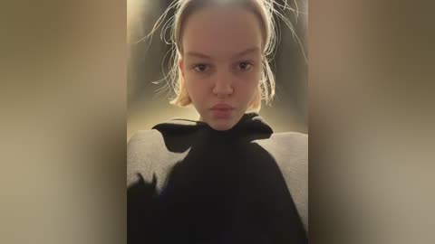 A video of a young woman with fair skin and blonde hair, wearing a black and white cow patterned sweater, pouting in a dimly lit hallway with blurred beige walls.