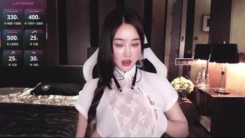 Video of a young East Asian woman in a revealing, sheer white maid outfit, with long black hair, in a dimly-lit bedroom.