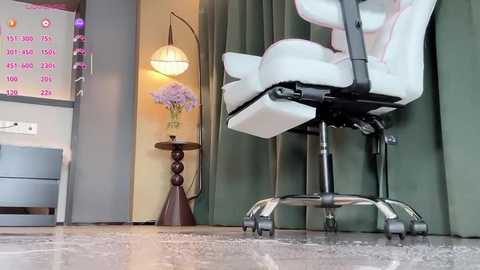 Video of a modern, minimalist office space featuring a white ergonomic chair, a small wooden side table with a flower vase, and a tall floor lamp with a curved design. The walls are painted in muted green and light beige, with a calendar displaying pink numbers on the left.