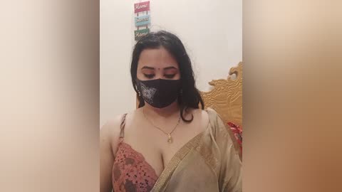 A video of a South Asian woman with medium skin tone, wearing a black face mask, a pink lace bra, and a beige saree. She stands in a narrow, beige-walled room, with a colorful box of condoms on her head.