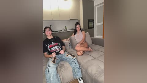 Video of a young couple lounging on a beige sectional sofa in a modern, minimalist living room. The man wears a black graphic T-shirt and jeans, while the woman is in a beige tank top and shorts.