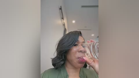 Video of a woman with shoulder-length, dark hair, wearing a green top, drinking from a clear glass, in a modern, white-walled room with a blurred background.