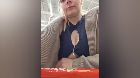 Video of a woman in a navy blue crop top with a keyhole cutout, wearing a beige cardigan, seated in a car. She has fair skin and blonde hair. Background shows a red can and a blurry, industrial ceiling.