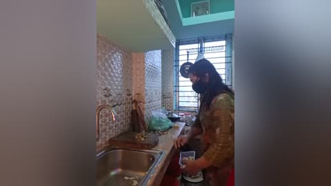 Video of a woman with long black hair and a face mask, washing dishes in a small, rustic kitchen with tiled walls and a window with white shutters.
