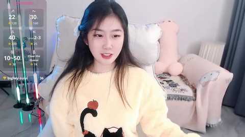 Video of an East Asian woman with long black hair, wearing a yellow sweater adorned with a cat graphic. Background features a plush animal bed and a virtual reality headset overlay.