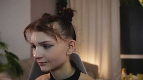 Video of a young woman with pale skin, dark hair in a bun, wearing black makeup, seated in a softly lit room with a window and green plant in the background.