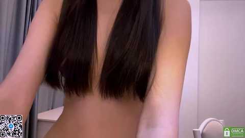 A video of a topless woman with long, straight black hair parted down the middle, standing indoors. The background features a white door and grey curtains.