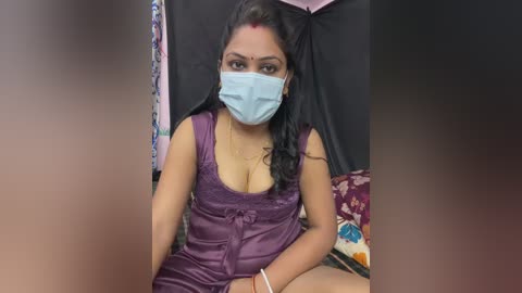 Media: Video of an Indian woman with medium-brown skin and long black hair, wearing a purple satin dress, face mask, and bindi, sitting in a tented room with colorful clothes hanging.
