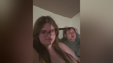 A video showing two young adults in a dimly lit room. A woman with long brown hair, glasses, and a gray top is in the foreground. A man with short hair, glasses, and a green shirt is behind her.