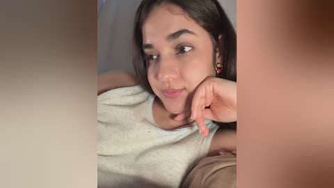 Video of a young South Asian woman with long dark hair, fair skin, and a slight smile, wearing a white sleeveless top. She is lying on a bed, with blurred, light-colored walls in the background.