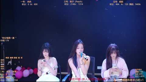 Video of three Asian women, two in white dresses, one in a pink jacket, singing into microphones against a dark stage with glowing pink balloons.