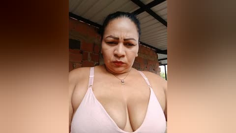 A video of a middle-aged woman with light brown skin and a round face, wearing a pink bra, standing in a rustic outdoor setting with a corrugated metal roof and brick wall background.