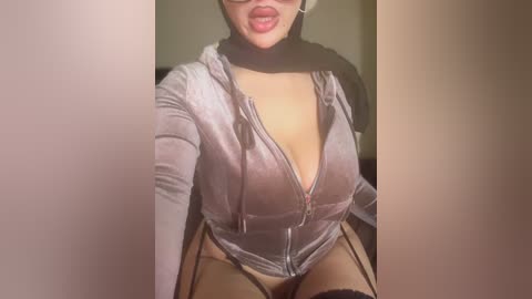 Video of a curvy, light-skinned woman with full lips, wearing a low-cut, grey velvet jacket and black lingerie, sitting in a dimly-lit room.
