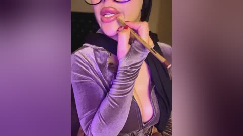 Video of a woman with fair skin, wearing a black hijab, glasses, and a low-cut, gray velvet top, holding a makeup brush near her lips, against a purple-hued background.