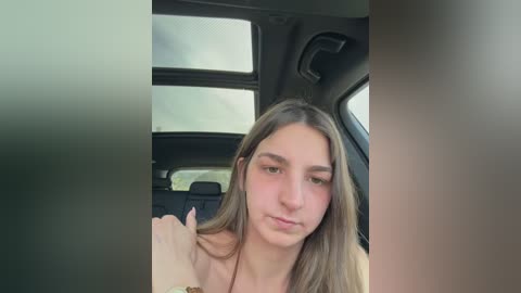 Video of a young woman with long, light brown hair, fair skin, and a neutral expression, sitting in the backseat of a car, looking out the window.