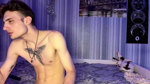 Video of a shirtless young man with a tattoo of wings on his chest, wearing a black necklace, standing in a dimly lit bedroom with a white bed, a black tapestry, and a small dog.