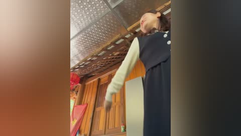A video of a young Asian woman in a black pinafore dress and white long-sleeve shirt, dancing vigorously with arms raised, set against a wooden-paneled room with a patterned ceiling and red decorations.