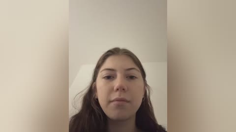 Video of a young Caucasian woman with straight, medium-length brown hair, fair skin, and neutral expression. She wears a white shirt, and the background is a plain, beige wall.