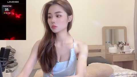 Video of an Asian woman with long brown hair, wearing a light blue satin robe, sitting on a bed, in a bedroom with a vanity mirror and makeup items.