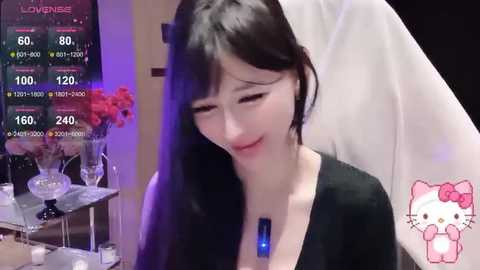 Media: A young Asian woman with long black hair and fair skin, wearing a black top, sits in a dimly lit room with a live stream overlay.