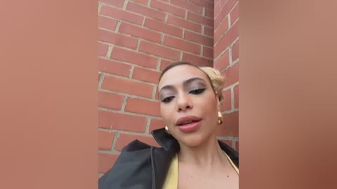 Video of a light-skinned woman with a medium build, dark hair styled in a bun, wearing a black leather jacket, gold hoop earrings, and heavy makeup, standing against a red brick wall.