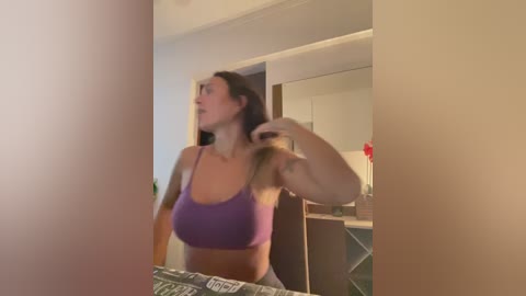 A video of a woman with light skin, long brown hair, and a medium build, wearing a purple sports bra, standing in a dimly lit bathroom. She's adjusting her hair with her left hand.