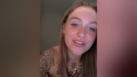 Video of a young woman with fair skin, brown hair, and a black tattoo sleeve on her left arm. She's smiling, leaning against a beige wall.