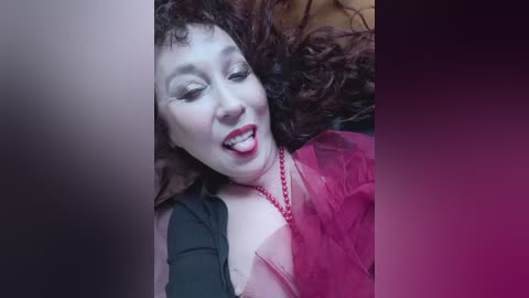 Media: Video of a woman with curly brown hair, wearing red lipstick, a black top, and a pink dress, lying on a bed, smiling broadly.