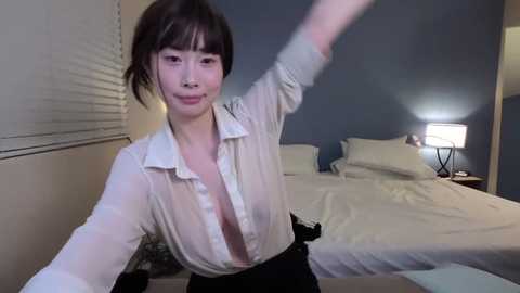 Video of a young Asian woman with short brown hair, wearing a sheer white blouse, black pants, and white socks, posing in a dimly lit bedroom with white bed, nightstand, and window blinds.