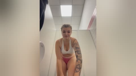 Video of a young, tattooed woman with light skin and blonde hair in a white sports bra and pink panties, walking down a narrow, brightly lit hallway.