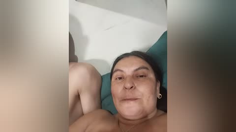 Video of a middle-aged woman with light skin, lying on a teal bedspread, wearing small hoop earrings, with a neutral expression.