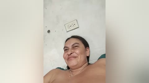 A video of a smiling, topless woman with medium skin tone, dark hair, and a necklace, standing in a plain, white-walled room with a light switch and outlet.