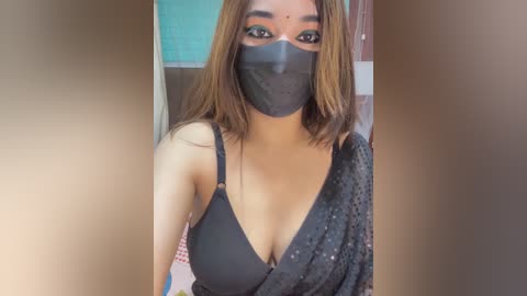 Video of a South Asian woman with medium skin tone, straight brown hair, and a black mask. She wears a black bra and a grey sari, standing indoors with a teal wall and wooden door background.