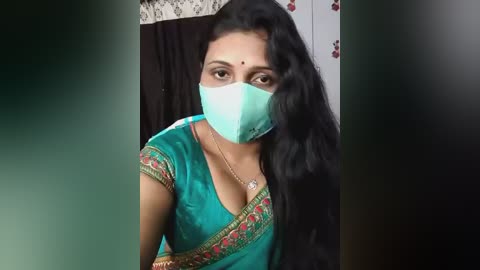 Video of a South Asian woman with long black hair, wearing a teal saree with gold embroidery, a green mask, and a gold necklace, standing in a room with floral wallpaper.