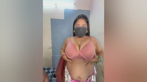 A video shows a curvy South Asian woman with medium skin tone, wearing a pink lace bra, black face mask, and floral skirt, posing indoors against a blue and white tiled wall.