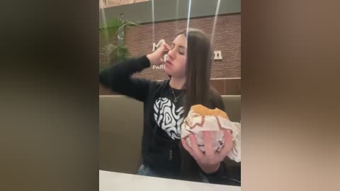 Video of a young woman with long black hair, wearing a black hoodie with a white design, licking a large ice cream cone with pink and yellow swirls, seated in a beige, cushioned booth.