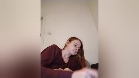 Video of a fair-skinned, red-haired woman in a burgundy sweater, leaning against a cream-colored wall with a power outlet visible in the background.