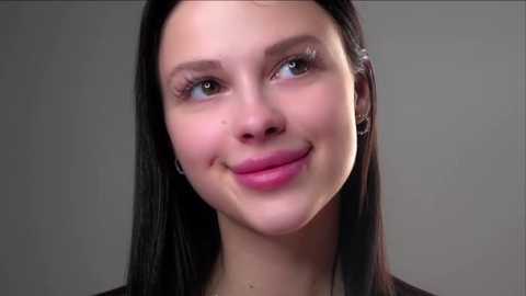 Video of a smiling young woman with light skin, straight dark hair, and blue eyes against a plain gray background. She wears a subtle makeup look and a dark top.