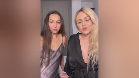 Video of two women standing side by side, one with long dark hair in a light gray satin camisole, the other with long blonde hair in a black silk robe.