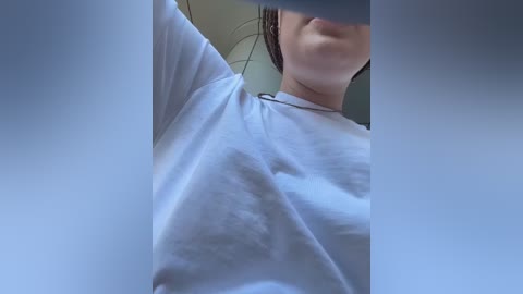 A close-up video of a woman wearing a white T-shirt, with her right arm raised, partially obscuring her face. Her dark hair is slightly wet and her skin is light. The background is blurry and neutral.