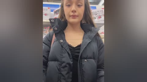 Video of a young Caucasian woman with light skin, brown hair, and a neutral expression, wearing a black puffy jacket over a black top, standing in a well-lit, modern grocery store aisle with shelves and product displays in the background.