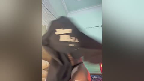 Video of a person wearing a black mask, holding a phone, in a dimly lit room with teal walls and white ceiling.